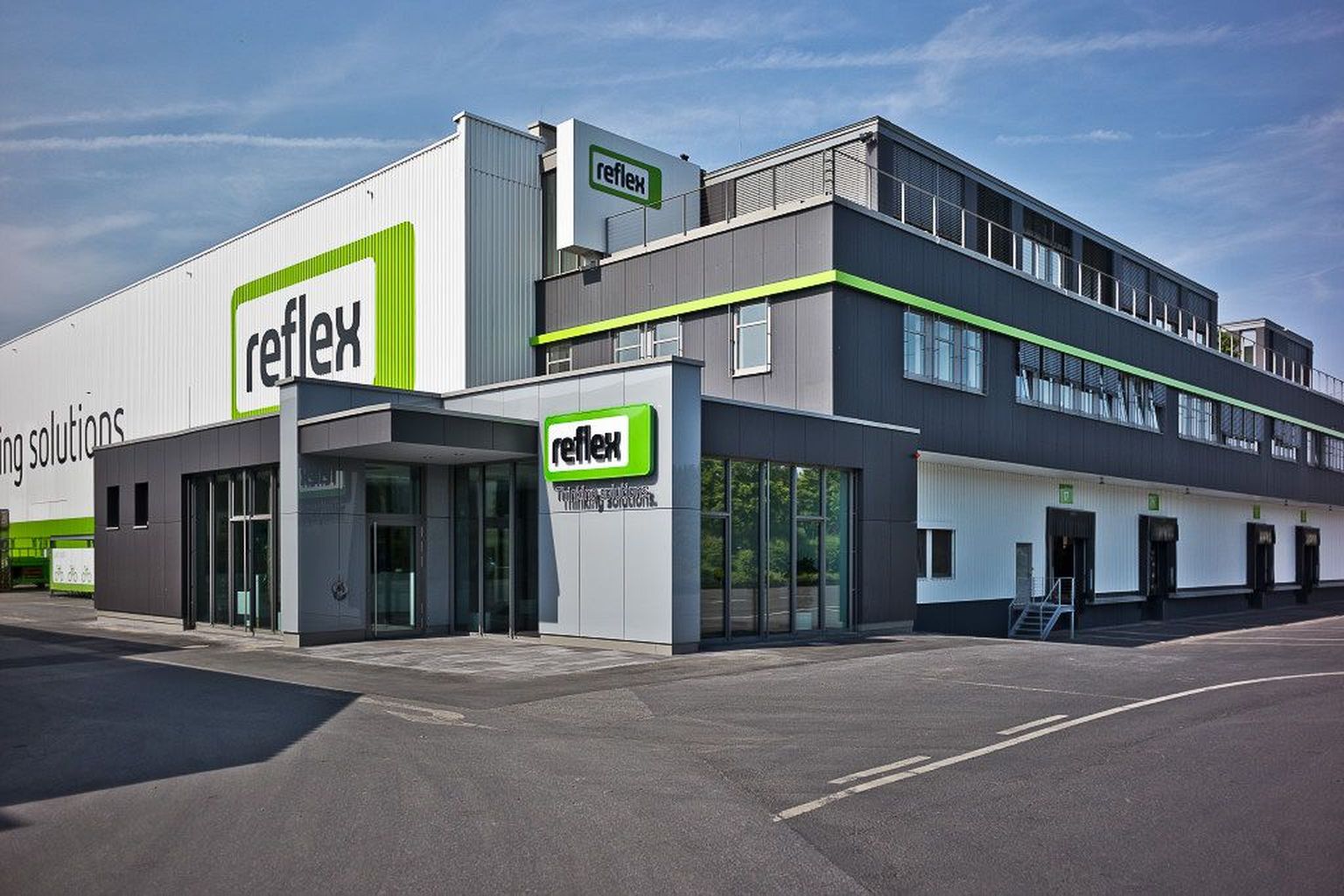 Modernes Industriegebäude mit „Reflex“-Beschriftung, grau-weißem Äußeren mit grünen Akzenten, großen Fenstern und einem angrenzenden Parkplatz.