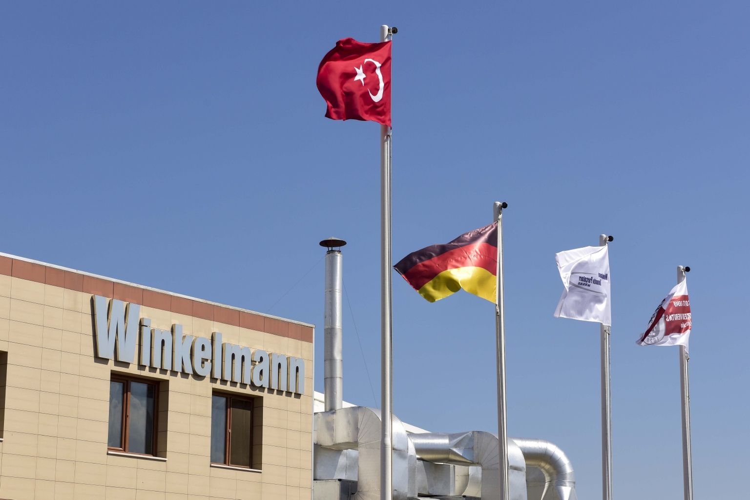 Winkelmann-Gebäude mit türkischen, deutschen und zwei Firmenflaggen an Masten vor einem strahlend blauen Himmel.