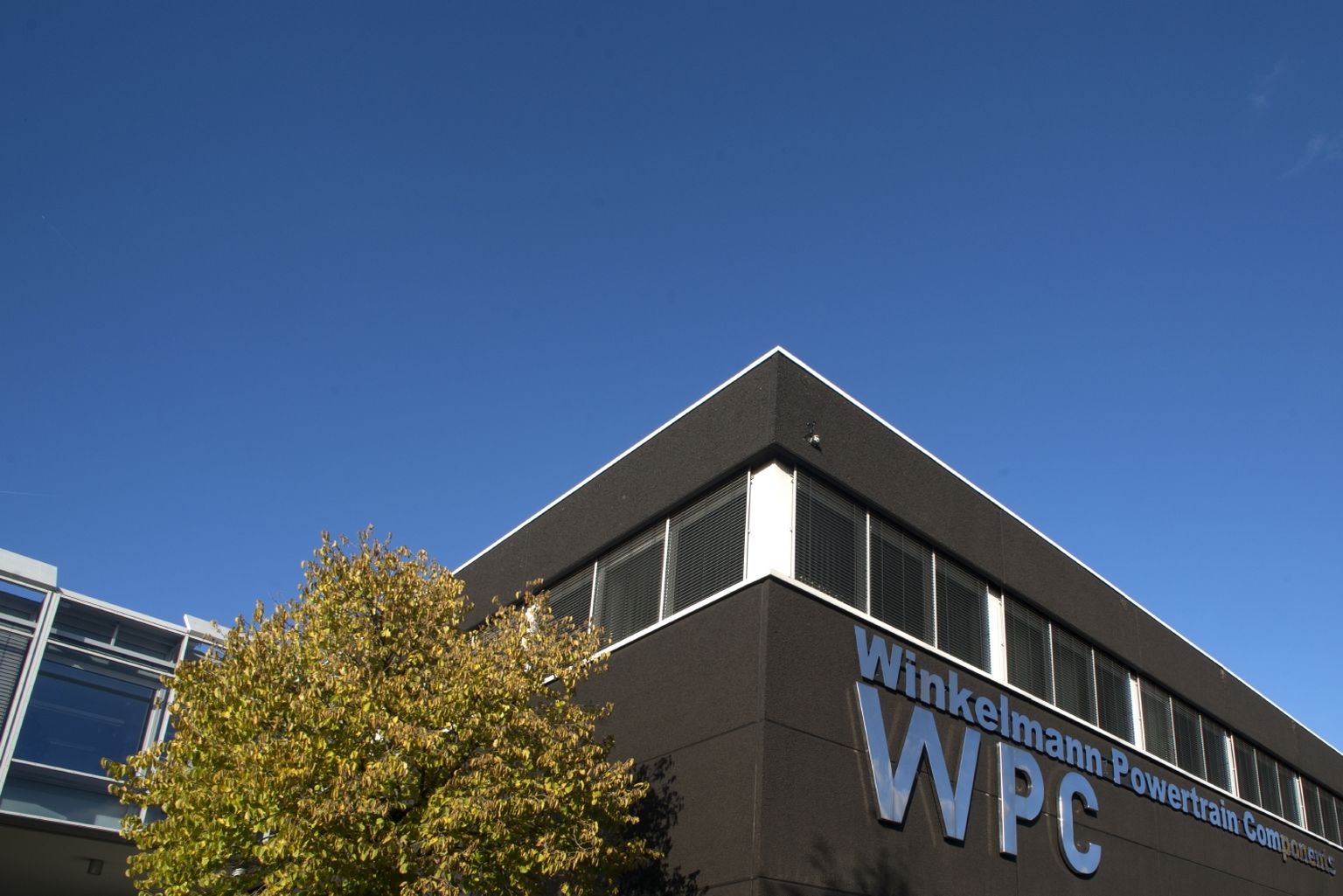 Modernes Gebäude mit der Aufschrift "Winkelmann Powertrain Components WPC“, klarer blauer Himmel und ein Baum auf der linken Seite.