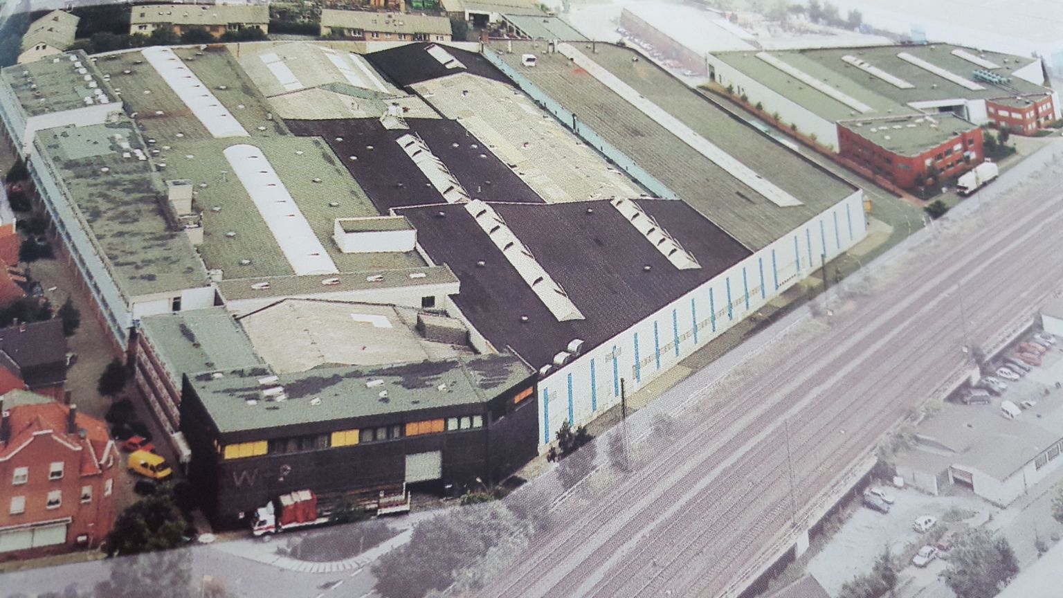 Luftaufnahme eines großen Industriekomplexes mit mehreren miteinander verbundenen Gebäuden, angrenzend an eine Bahnlinie und umgeben von Bäumen und Straßen.
