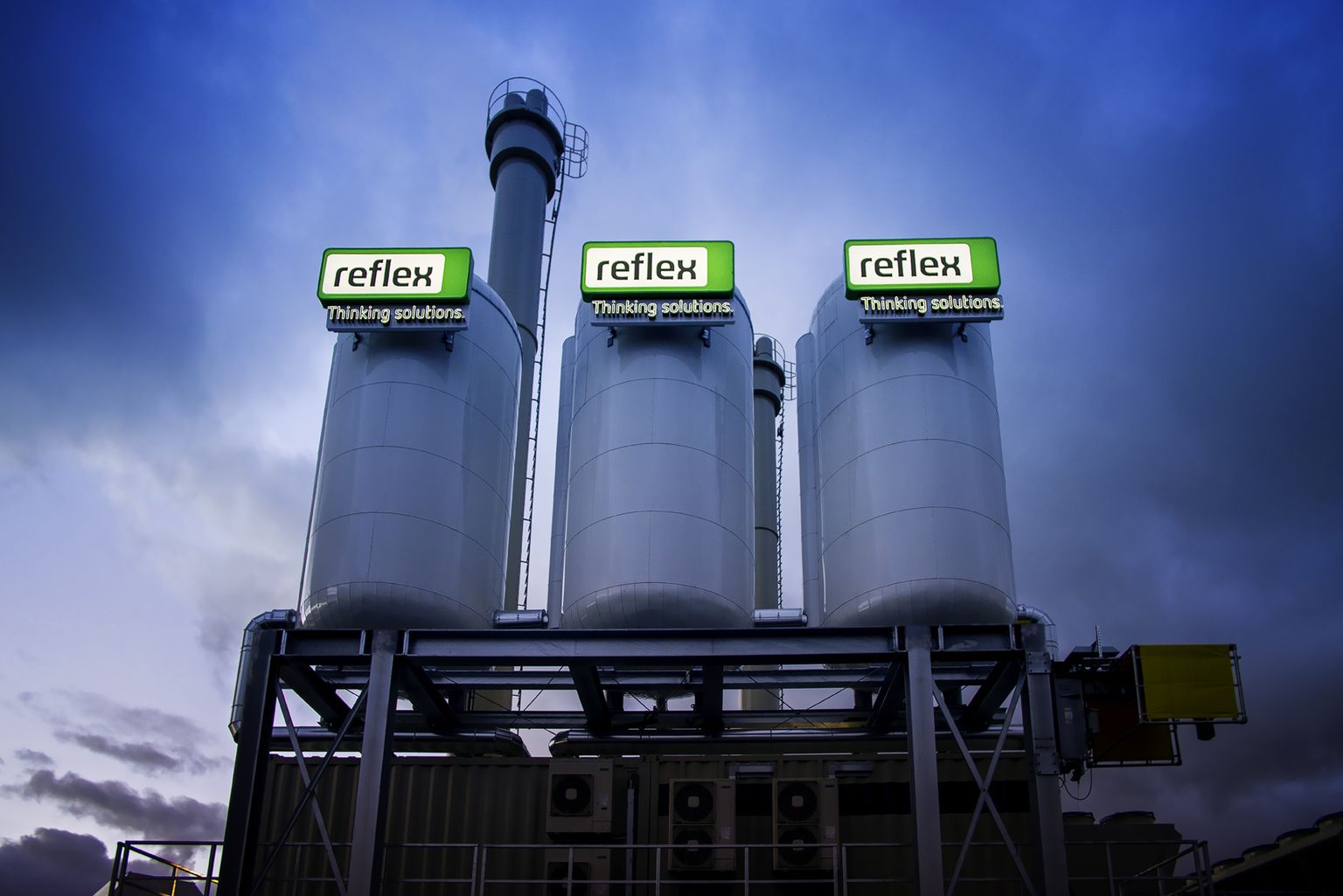 Drei große Industrietanks mit „Reflex“-Schildern auf der Oberseite vor einem wolkenverhangenen Himmel, mit dem Slogan „Thinking solutions“.