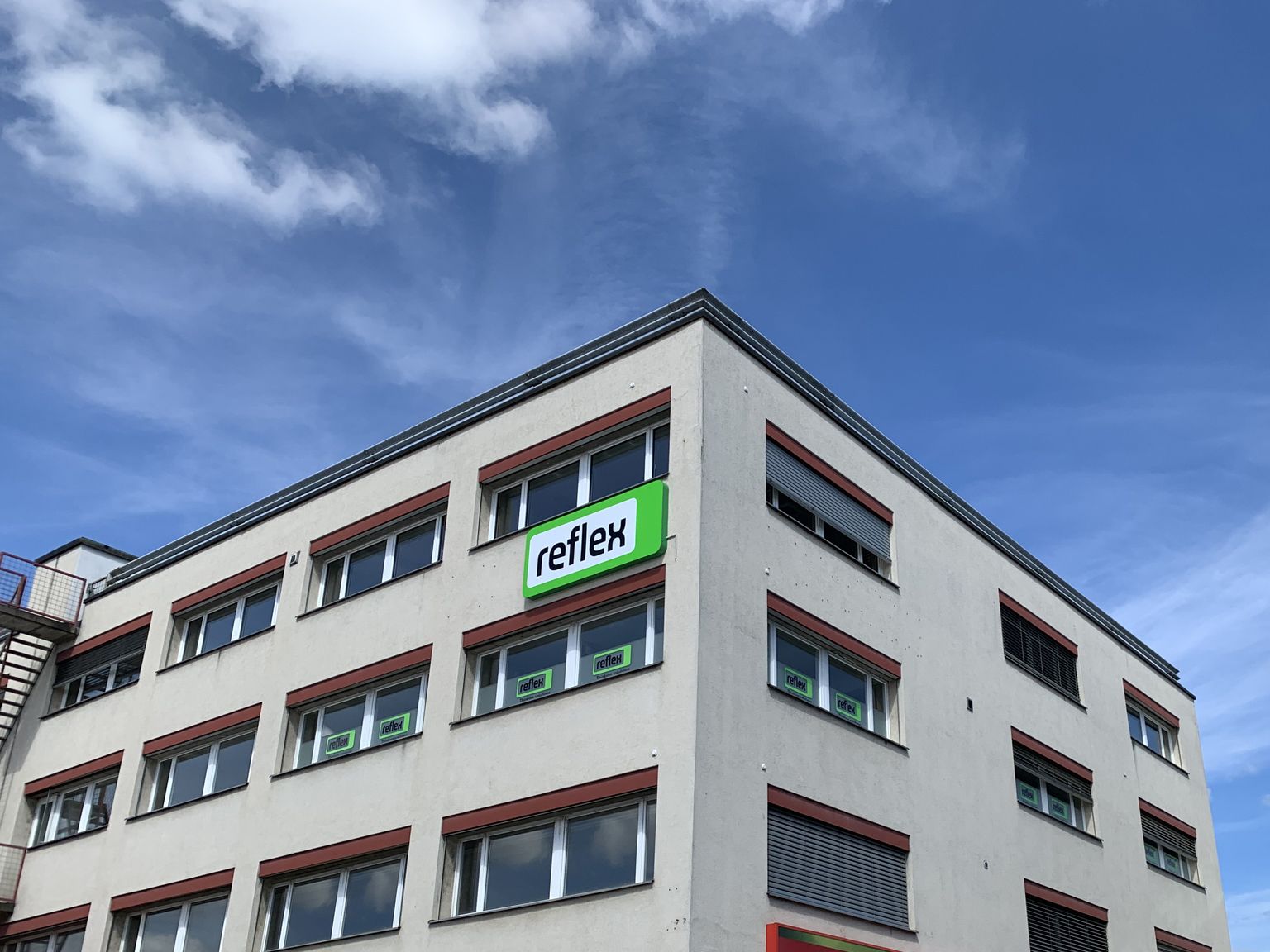 Ein modernes, mehrstöckiges Gebäude mit einem Schild mit der Aufschrift „reflex“ unter einem blauen Himmel mit Wolkenfetzen.