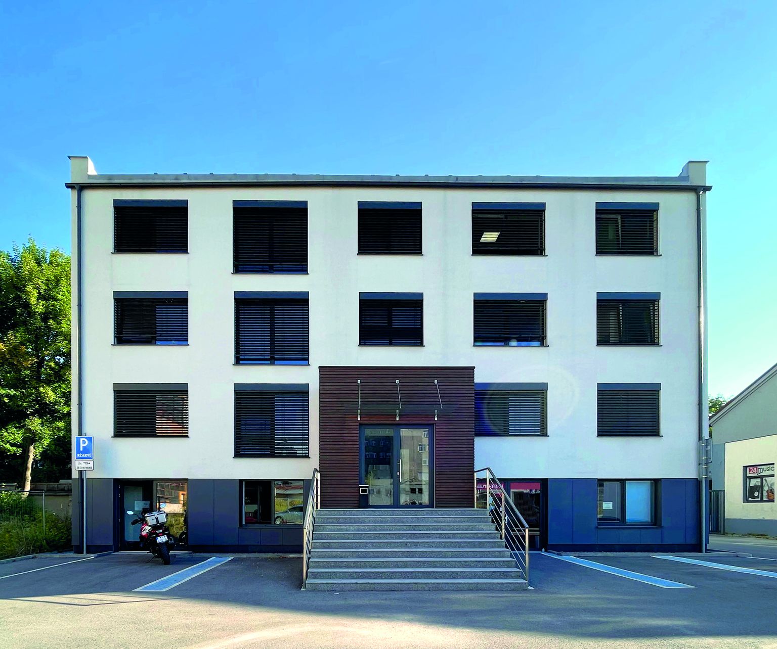 Ein modernes dreistöckiges Gebäude mit einer weißen Fassade, großen Fenstern und einer zentralen Treppe, die zum Haupteingang führt.