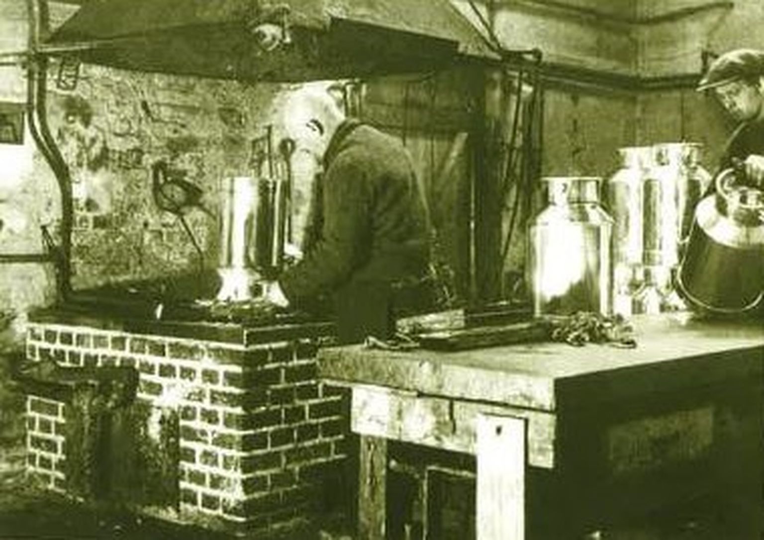 Zwei Männer arbeiten in einer alten Werkstatt und stellen große Metallbehälter auf einer gemauerten Schmiede her. Die Kulisse hat ein altmodisches, industrielles Flair.