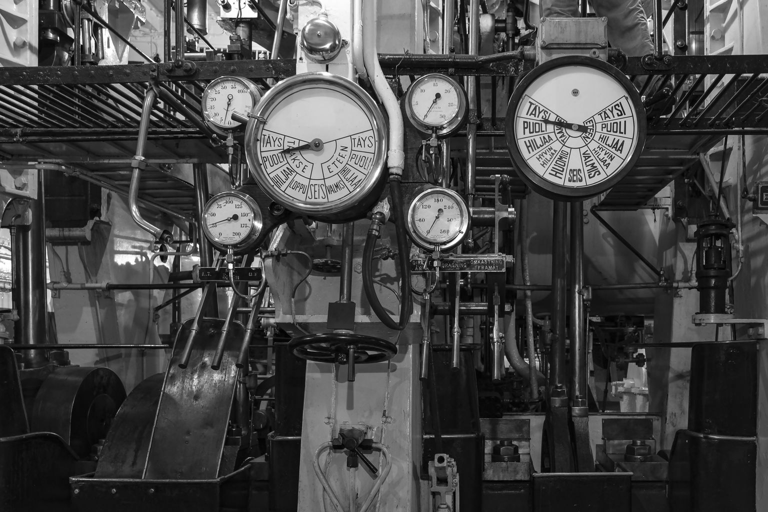 Schwarz-Weiß-Bild einer komplexen industriellen Schalttafel mit mehreren Zifferblättern, Messgeräten und Hebeln in einer mechanischen Umgebung.