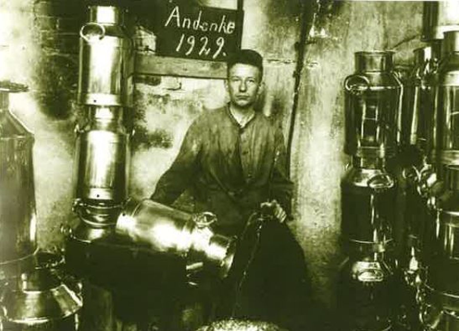 Mann in einer Werkstatt, umgeben von glänzenden Metallbehältern, an der Wand ein Schild mit der Aufschrift „Andenke 1929“.