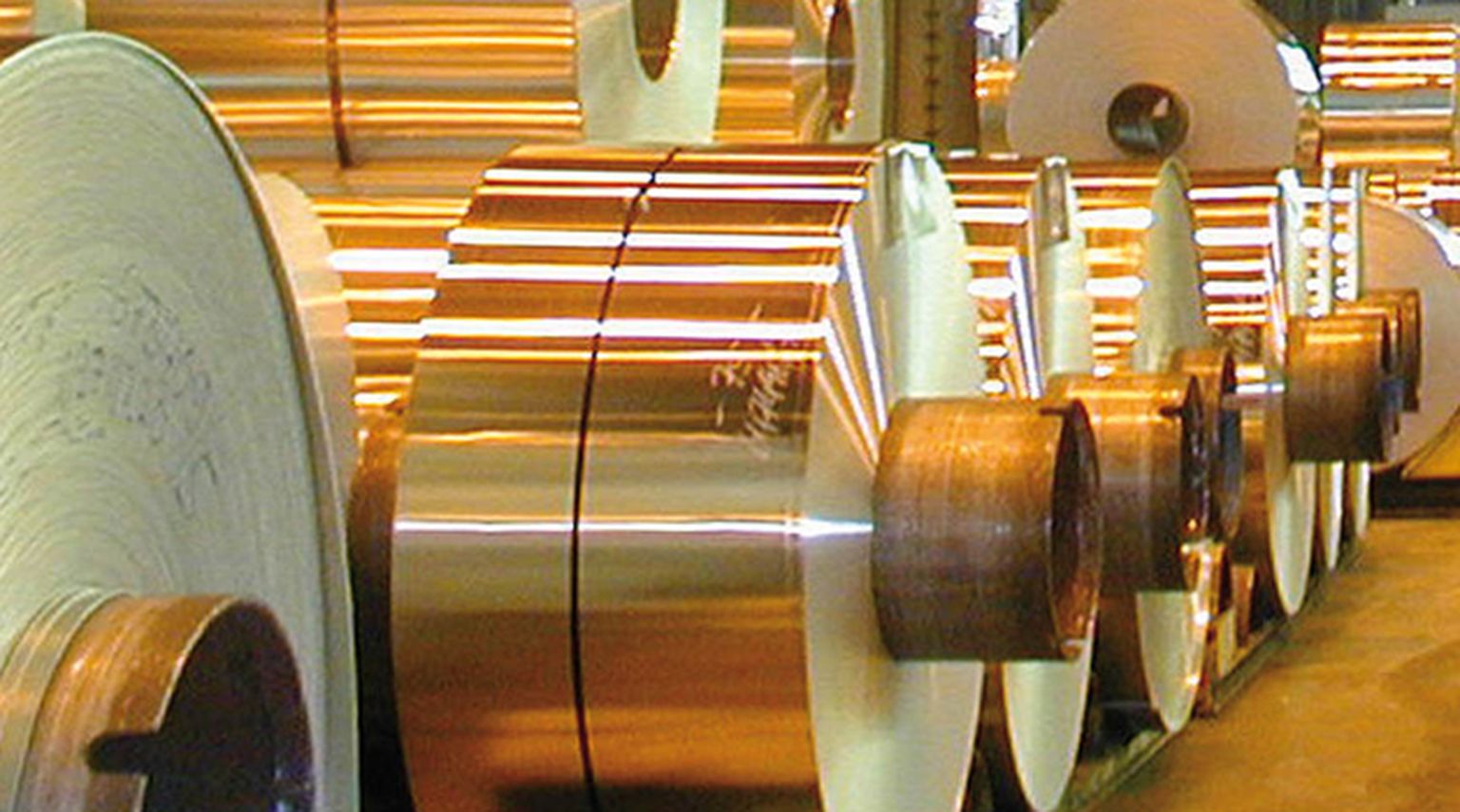 Große Rollen glänzender Kupferbleche sind in einer industriellen Umgebung aufgereiht, reflektieren das Licht und bringen ihre metallische Textur zur Geltung.