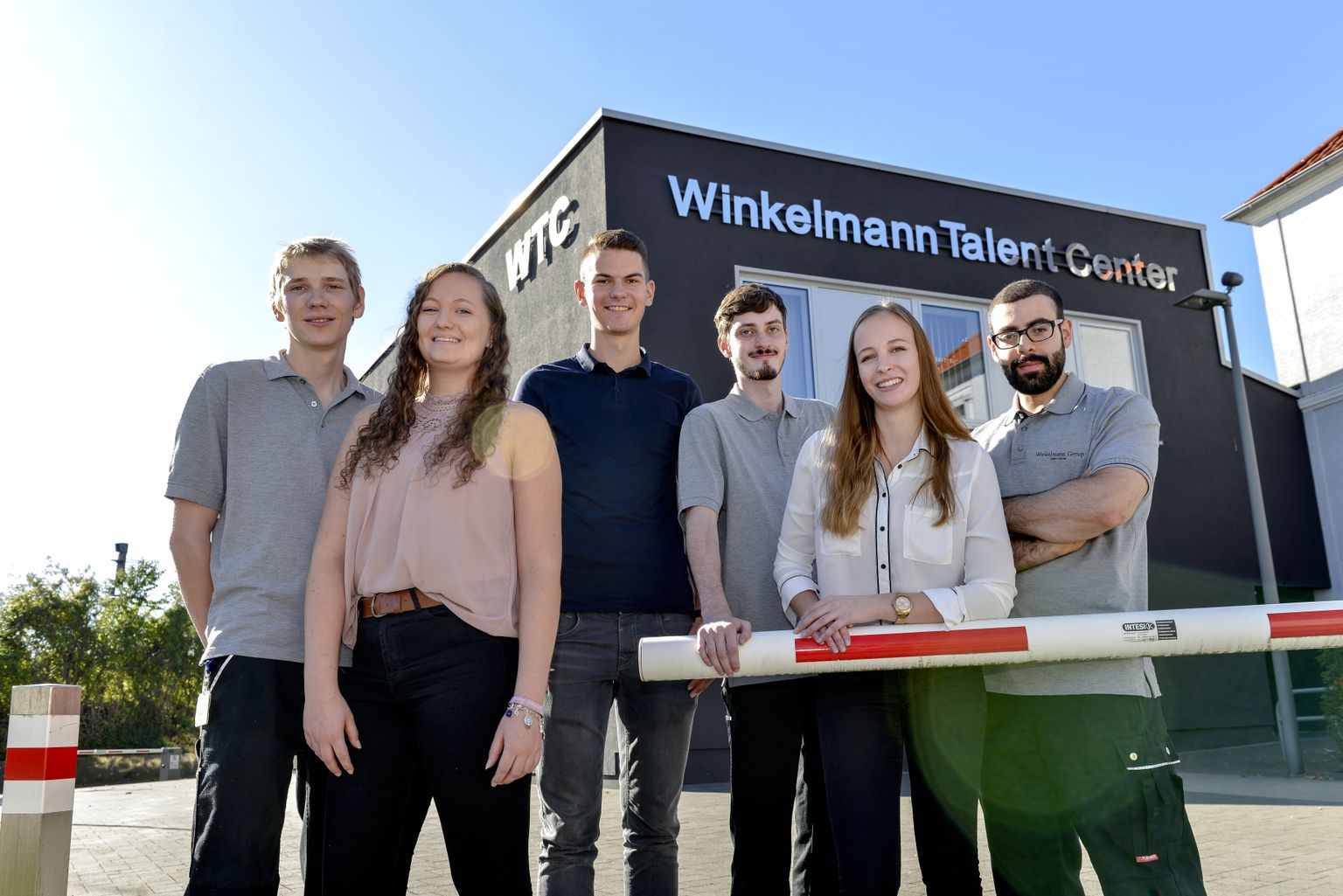 Sechs Personen stehen vor einem Gebäude mit der Aufschrift „Winkelmann Talent Center“, lächeln und posieren für das Foto.