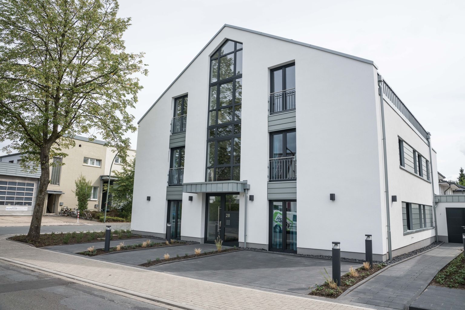 Modernes weißes Gebäude mit großen Fenstern und minimaler Landschaftsgestaltung. Das Hotel liegt an einer ruhigen Straße mit einem Baum und gepflasterten Parkplatz.