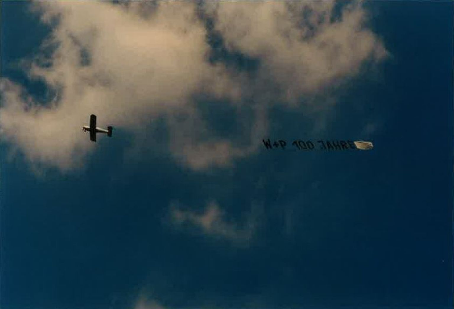 Ein kleines Flugzeug fliegt bei teilweise bewölktem Himmel und schleppt ein Banner mit der Aufschrift „W+P 100 JAHRE“.
