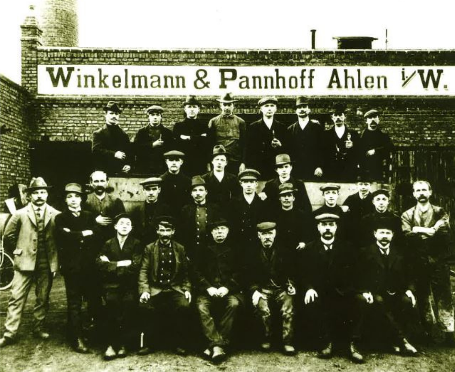 Ein altes Gruppenfoto von Männern in Arbeitskleidung, die vor einem Backsteingebäude mit der Aufschrift „Winkelmann & Pannhoff Ahlen“ posieren.